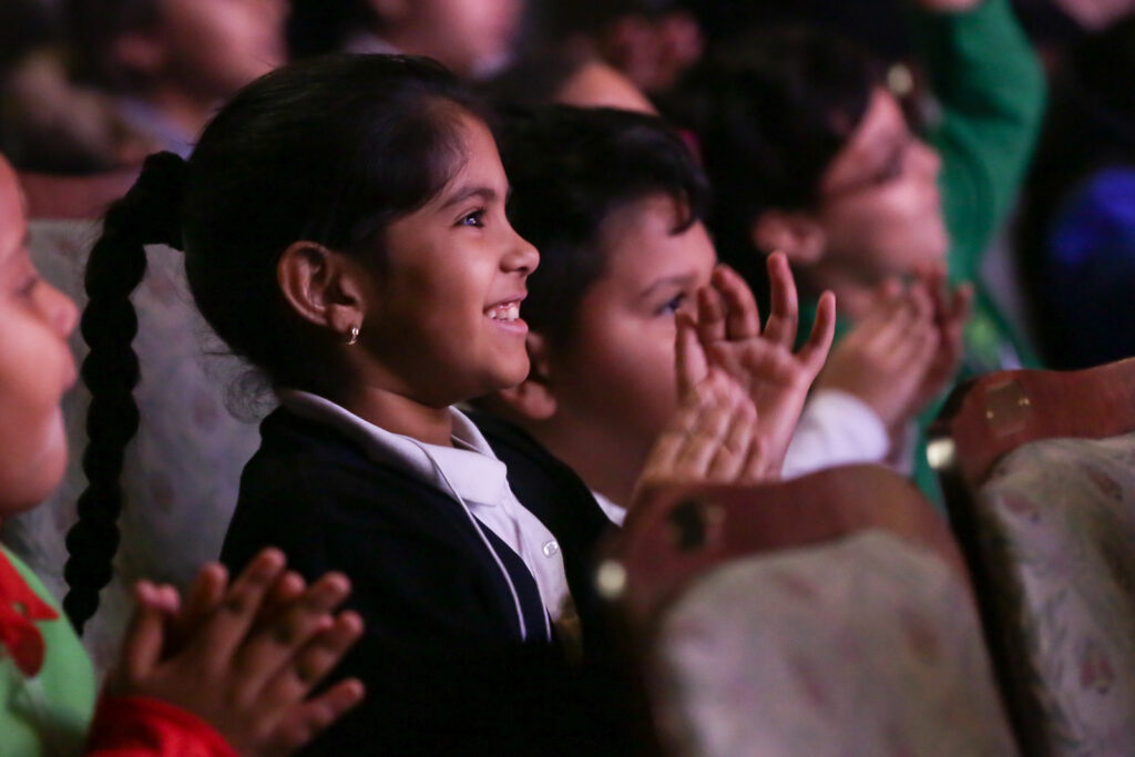 Give-A-Kid-A-Seat-Donation-Page-1024×683 A young girl reacts to a performance with a smile and hands clapping.
