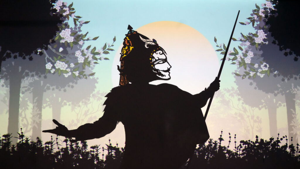 A performer wears a headpiece that casts a shadow of a face against a projection of trees and the sun.