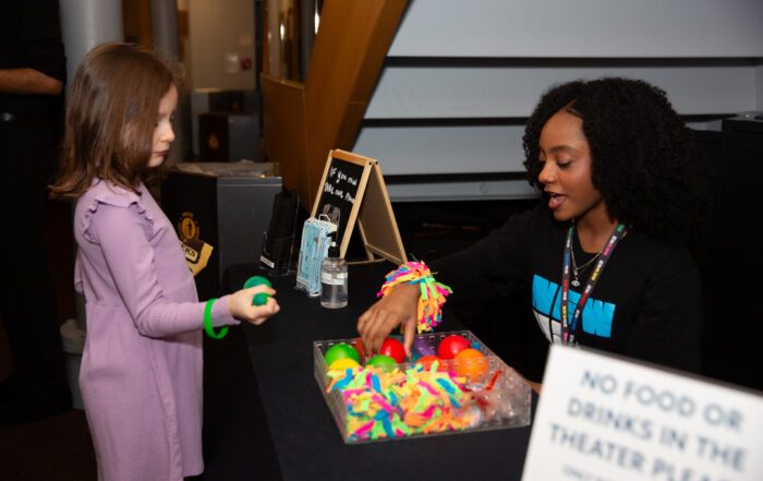 A photo of two ushers at the Accessibility Table giving fidget toys to young patrons.