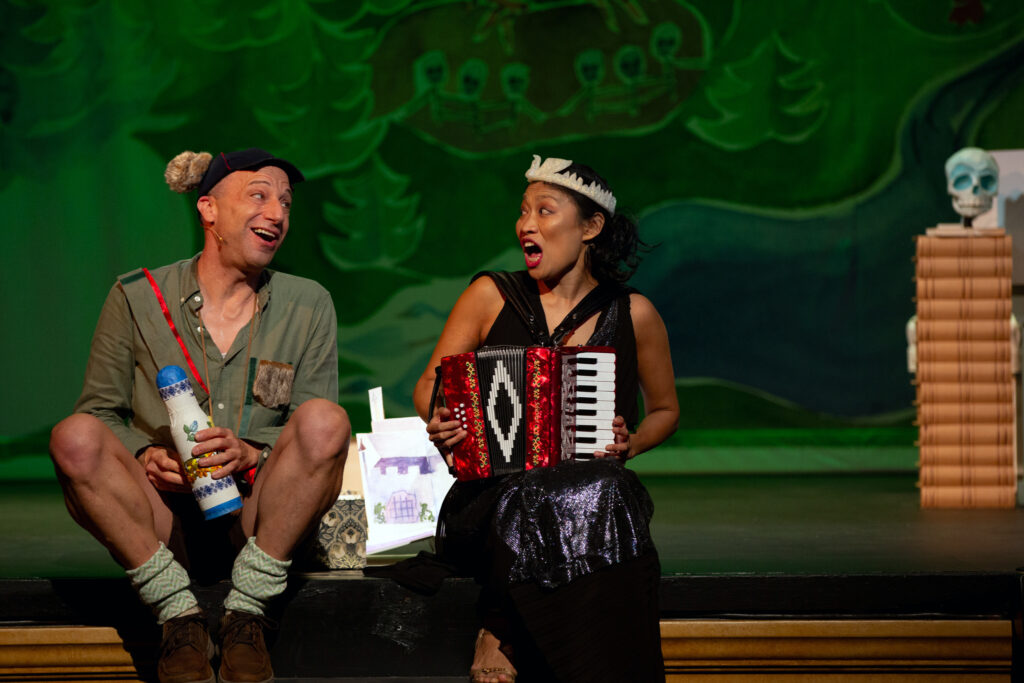 Anton Dudley and Faye Chiao sit on the edge of the stage singing while Faye plays accordion.