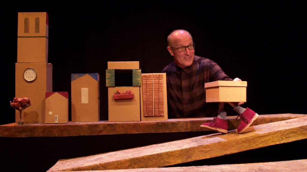 A man in glasses and a striped maroon shirt stands next to a row of five cardboard boxes built to look like a street and moves a puppet of a shoebox with a pair of ankles, white socks and red shoes down a ramp in front of them.