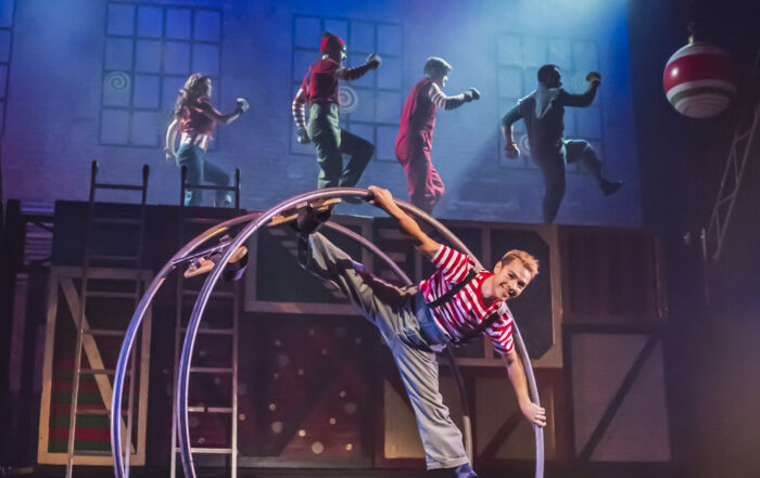A man wearing a stripped shirt and grey overalls performing with a Cyr wheel.