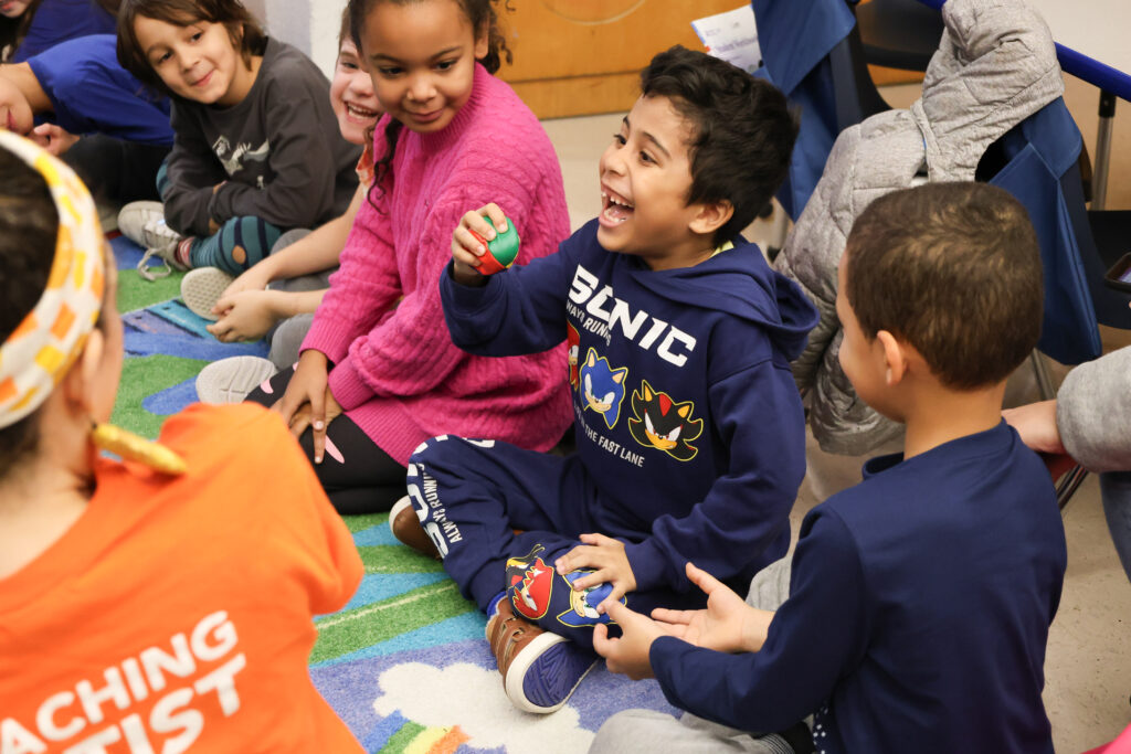 A group of kids sitting on a rug with one of the kids holding a small bean bag.