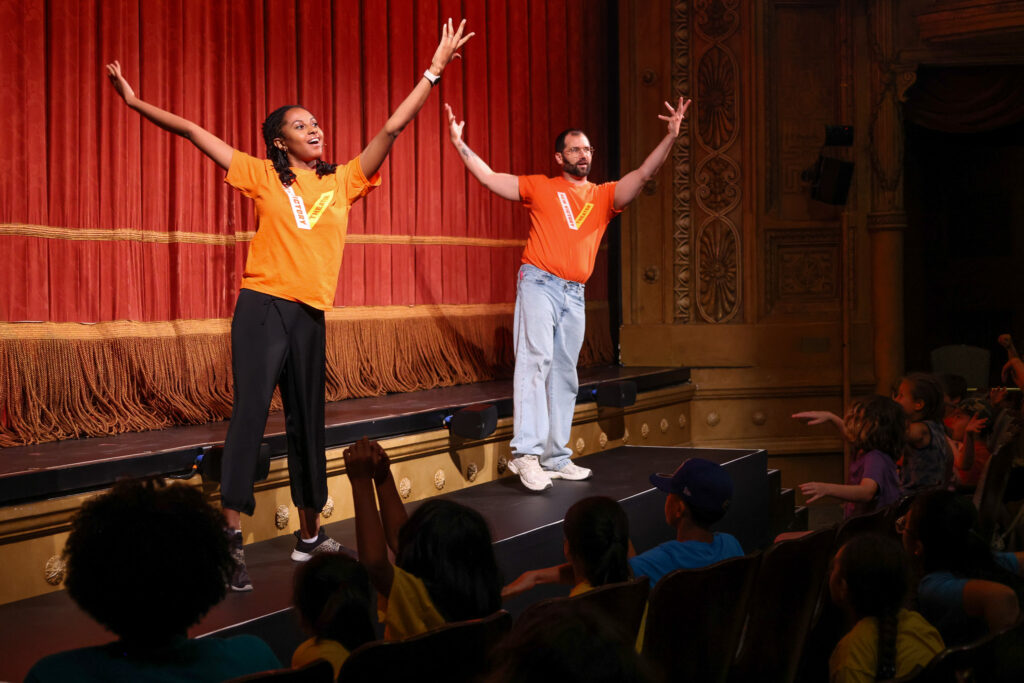 Two New Victory teaching artist on stage performing with their hands up.