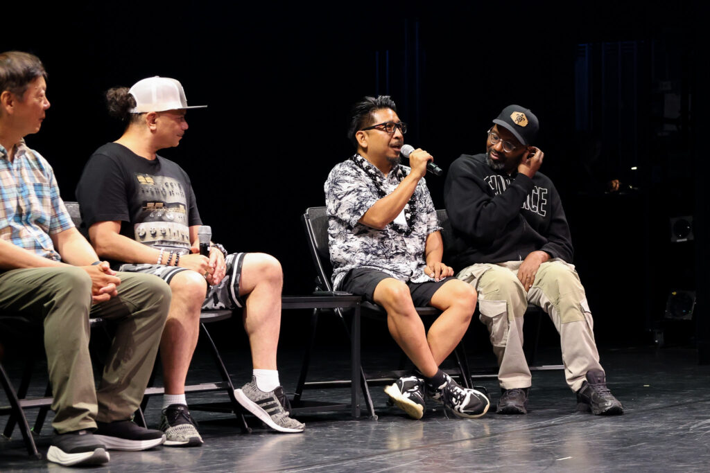 Four men on stage sitting on chairs with one of the men speaking into a microphone.